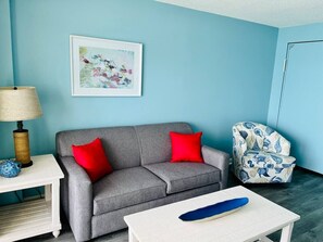 A room with a grey sofa, red cushions, a white table, a blue chair, and a framed picture on the wall. - Paradise by the Sea. Oceanfront. (Myrtle Beach)
