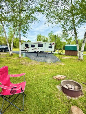 Un camping con una caravana, una silla roja y una fogata.