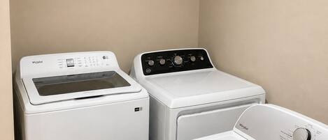 A laundry room with a front-loading washer, a top-loading dryer, and a shelf with a laundry basket and a potted plant.