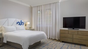 Ett hotellrum med en säng, en tv, ett nattduksbord, en byrå och ett fönster med tunna, genomskinliga gardiner.