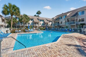 A swimming pool with lounge chairs and palm trees, surrounded by multi-story buildings. - Courtside 67 - Your Perfect Hilton Head Island Getaway (Hilton Head Island)