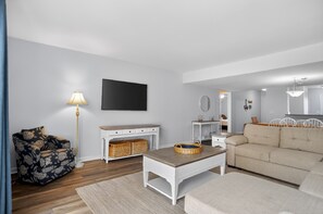 A modern living room with a sofa, a coffee table, a lamp, a television, and a dining area with chairs. - Courtside 67 - Your Perfect Hilton Head Island Getaway (Hilton Head Island)