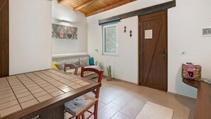 Een kamer met houten plafond, een eettafel met stoelen, een bank, een raam met gordijnen en een deur met een bordje.
