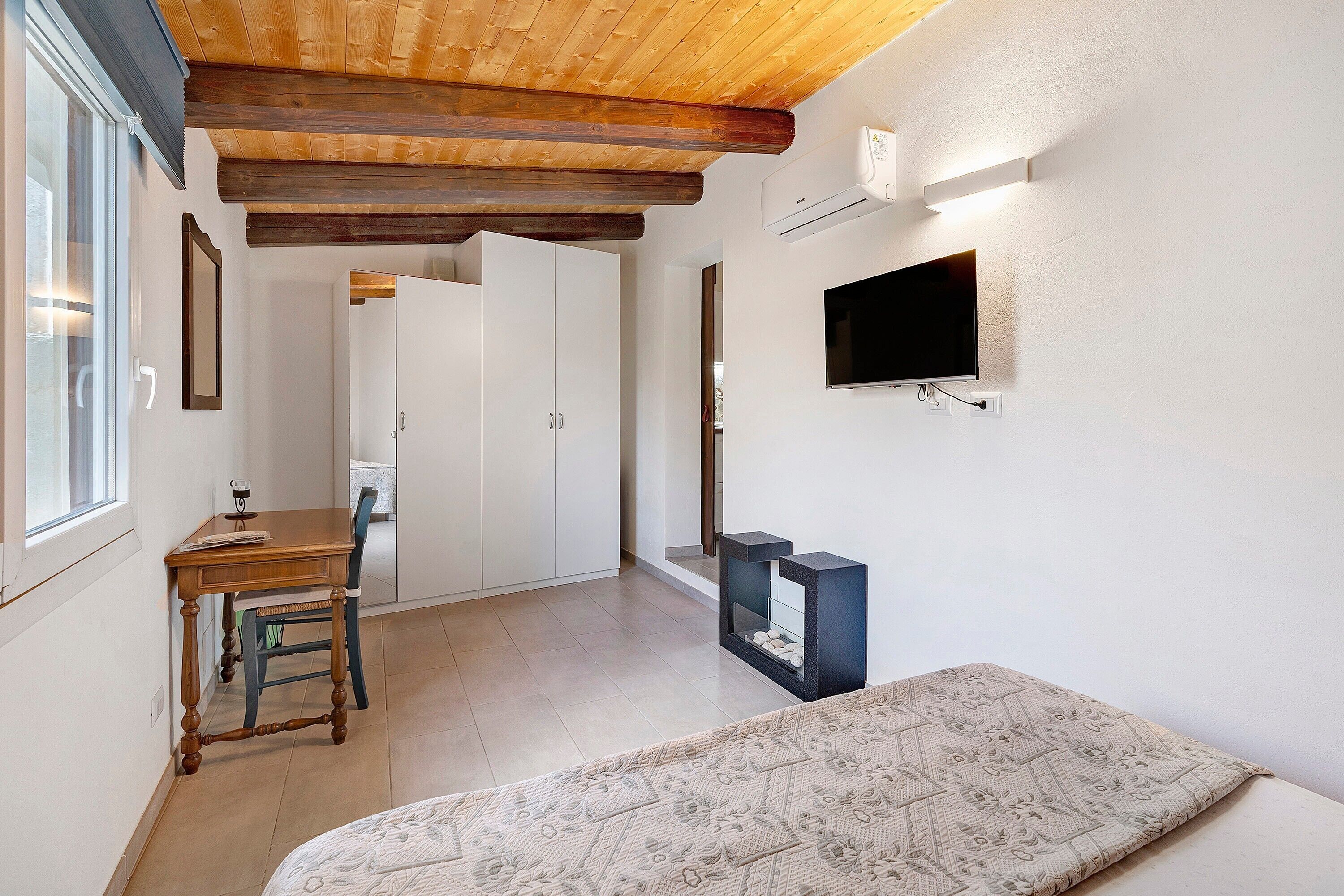 A room with a wooden ceiling, a television mounted on the wall, a bed with a patterned bedspread, and a desk with a chair.
