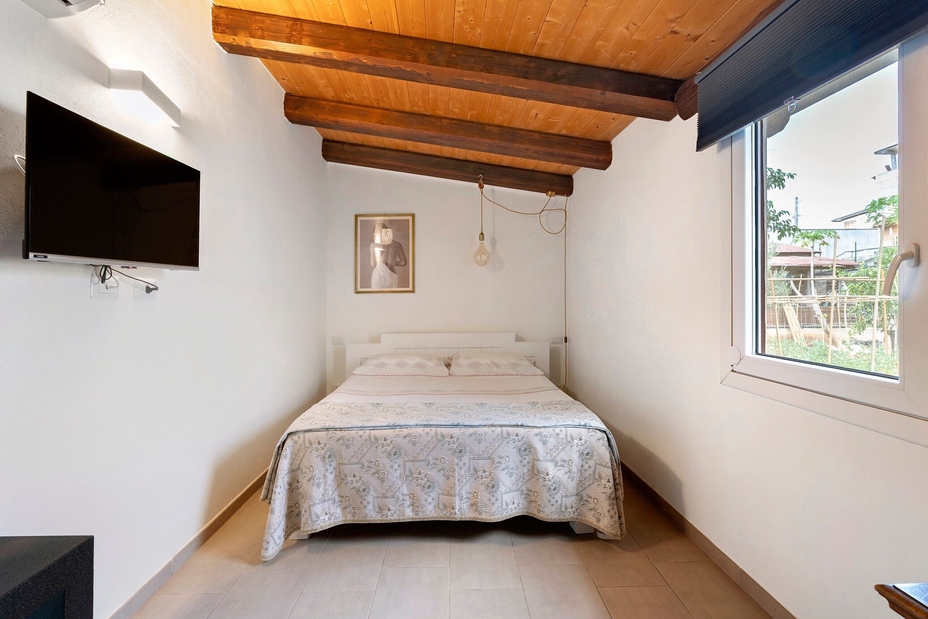 Ein Schlafzimmer mit einem Bett, einem an der Wand befestigten Fernseher, einem gerahmten Bild und einem Fenster.