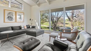 Ein modernes Wohnzimmer mit einem Ecksofa, einem Lederhocker, einem runden Couchtisch und einem Sessel mit Webkissen. Große Fenster bieten Blick ins Freie.