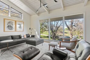 A modern living room with a sectional sofa, a leather ottoman, a round coffee table, and a chair with a woven backrest. Large windows provide a view of the outdoors. - Aalto Modern - Spacious, 5-bed MCM - A Luxury KY Derby Vacation! (Louisville)