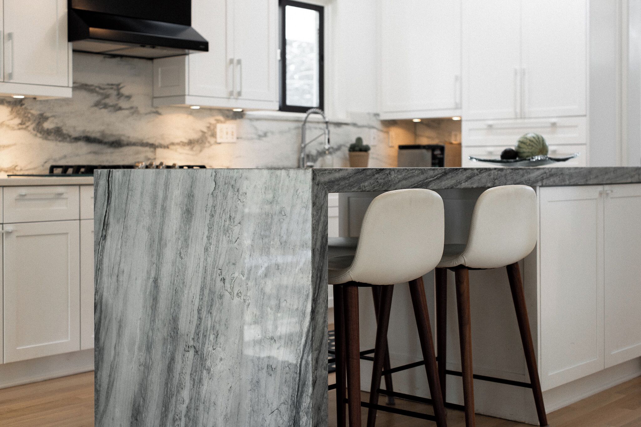 Cocina con encimeras de mármol, gabinetes blancos y dos taburetes de bar.