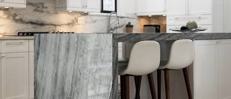 Cocina con encimeras de mármol, gabinetes blancos y dos taburetes de bar.
