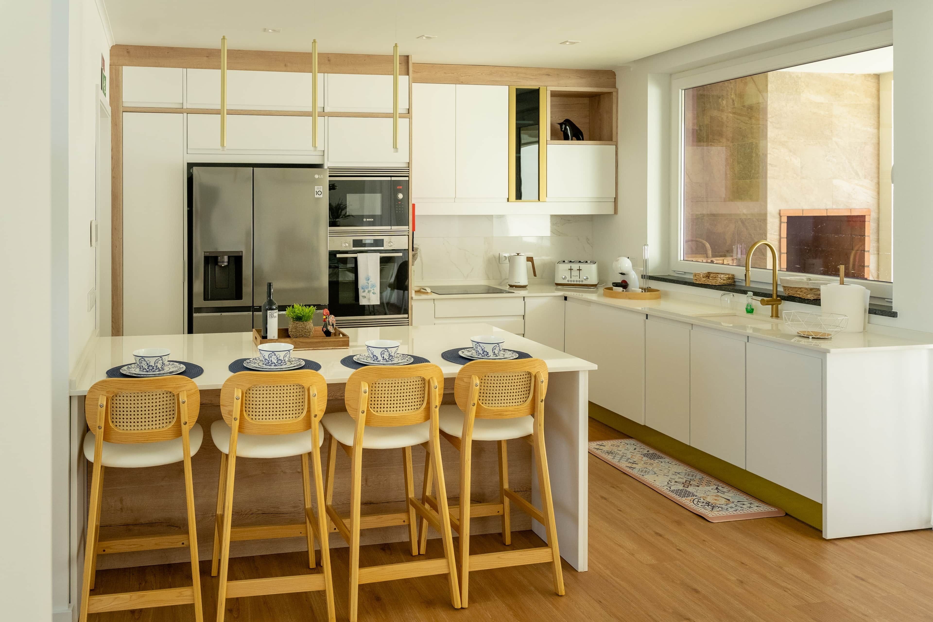 Cocina moderna con una isla central, cuatro sillas de madera, horno empotrado, refrigerador y ventana con vistas.