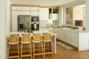 Une cuisine moderne dotée d’un îlot central, de quatre chaises en bois, d’un four encastré, d’un réfrigérateur et d’une fenêtre offrant une vue agréable.