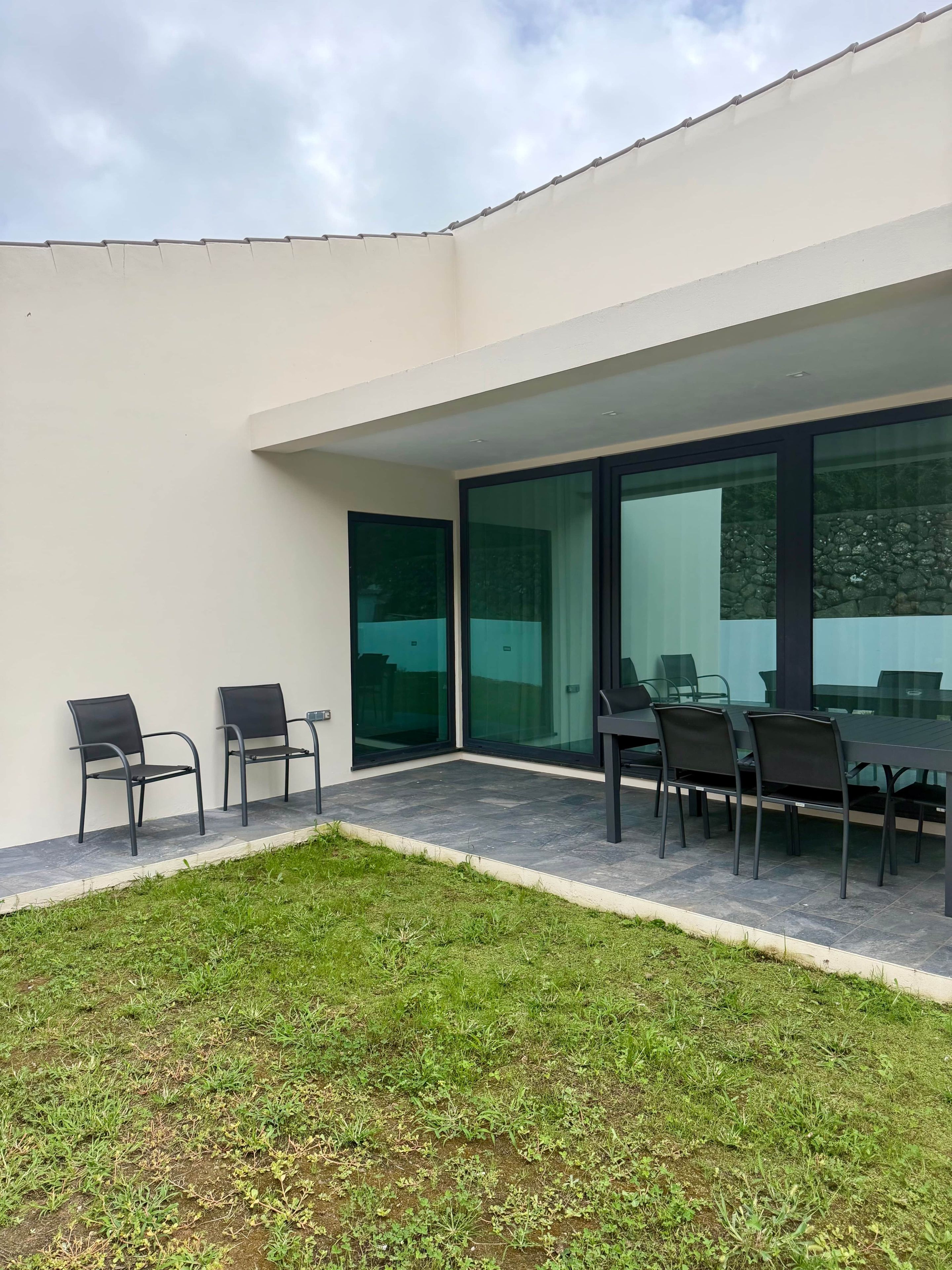 Uma área de estar moderna ao ar livre, com mesa e cadeiras, gramado e um edifício com grandes janelas de vidro.