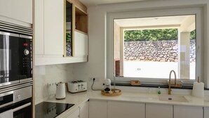 A modern kitchen with a built-in oven, a coffee maker, and a window with a view of a stone wall and greenery. - Casa das Hortênsias (Nossa Senhora do Pilar Terceira)