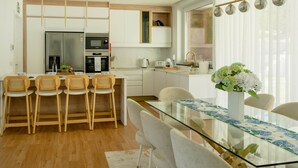 A modern kitchen with white cabinets, a stainless steel refrigerator, and a dining area with a glass table and chairs. - Casa das Hortênsias (Nossa Senhora do Pilar Terceira)