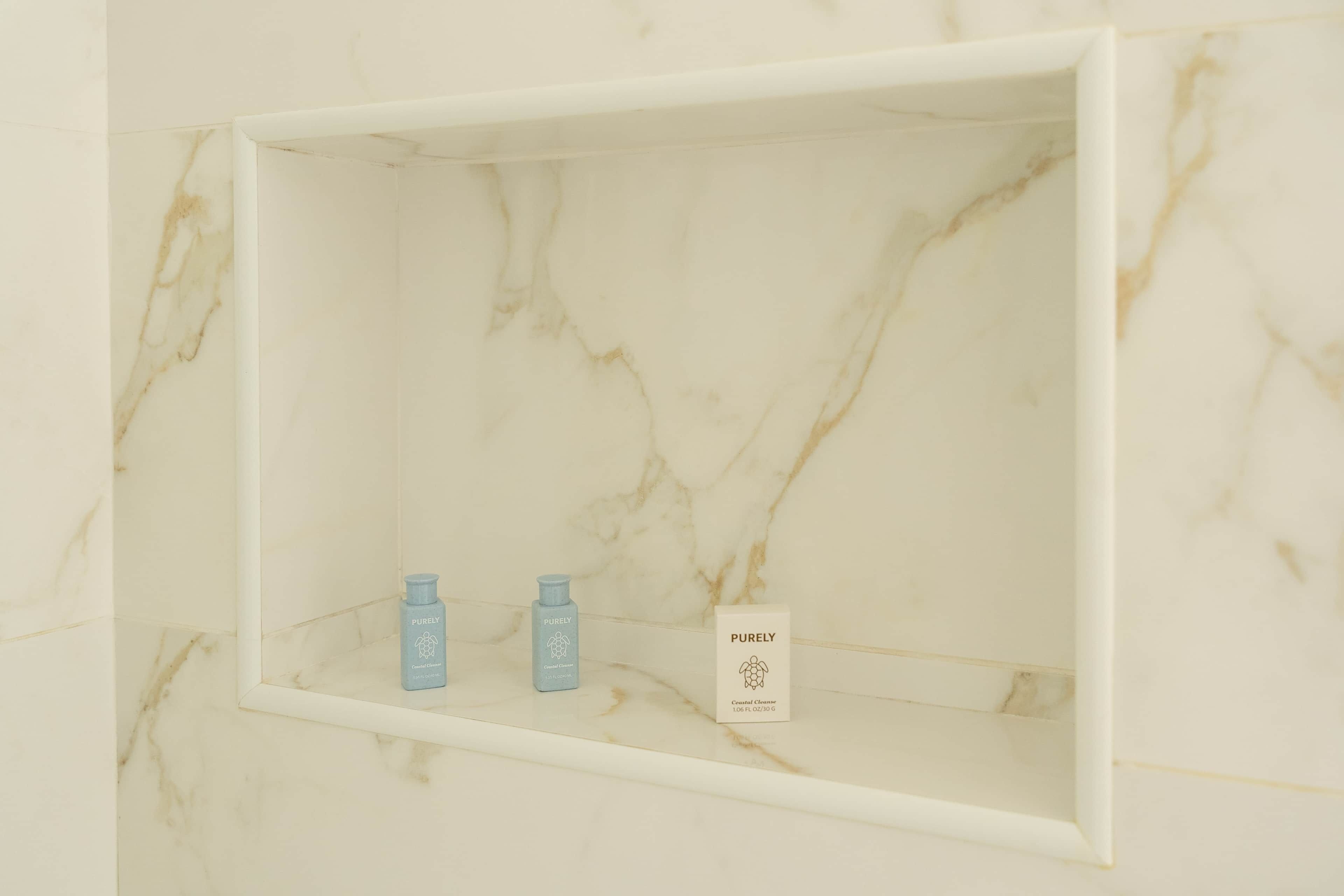 Una prateleira de bany amb paret de marbre, amb dues amples de Purely Central Cleanser i una caixa de Purely Coastal Cleanse.