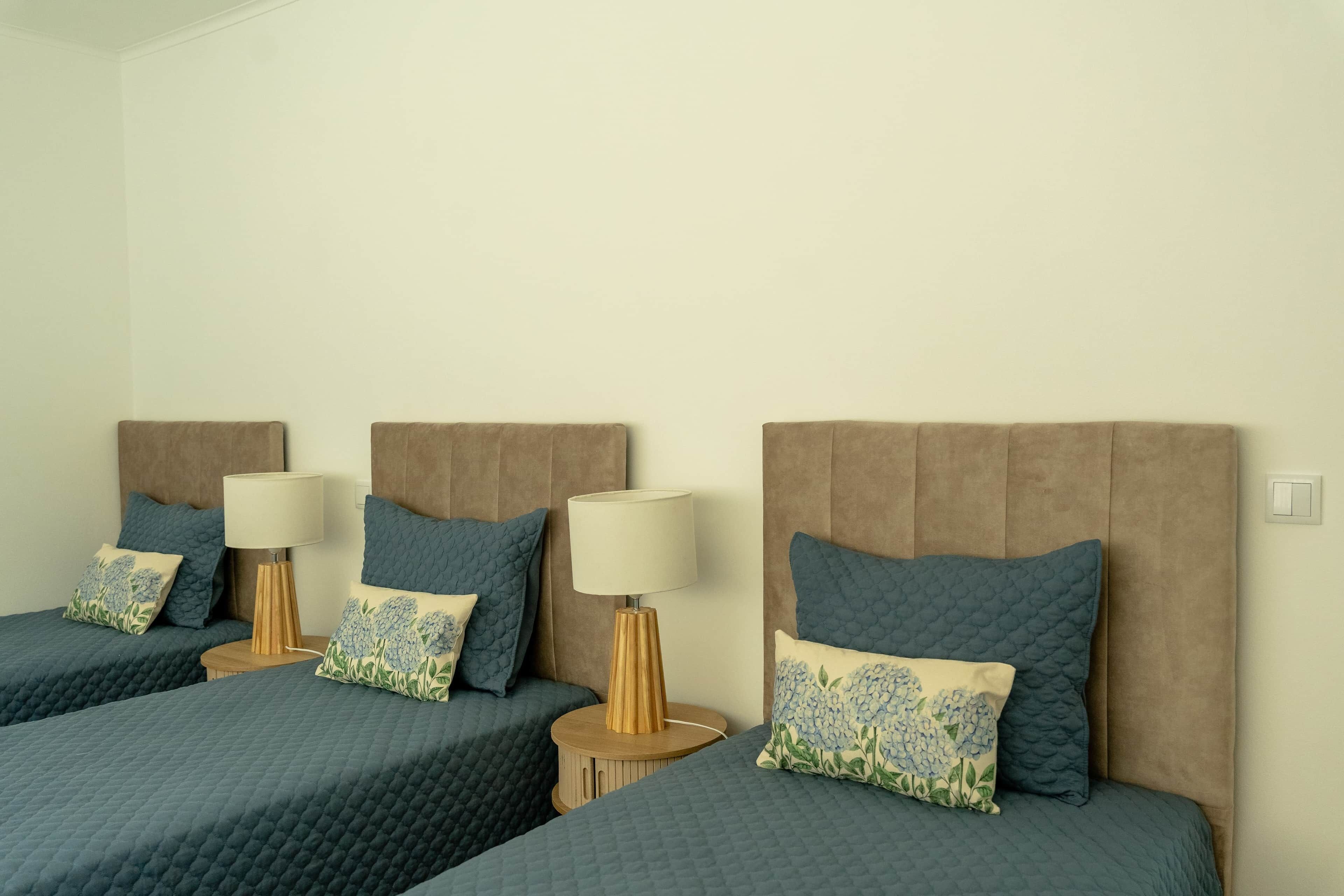 Habitación de hotel con tres camas, cada una con cabecera, mesitas de noche, lámparas y almohadas decorativas.