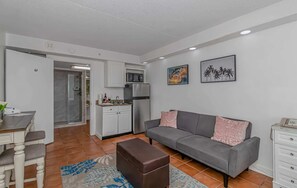 Un salon moderne avec un canapé gris, un pouf marron et un coin repas comprenant une table et des chaises blanches.
