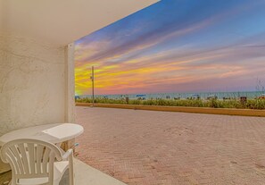 Ein gepflasterter Außenbereich mit Blick auf Strand und Meer, ein weißer Tisch und ein weißer Stuhl.