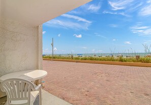Eine Terrasse mit einem weißen Kunststoffstuhl und einem Tisch, mit Blick auf einen Strand mit Sonnenschirmen und Menschen.