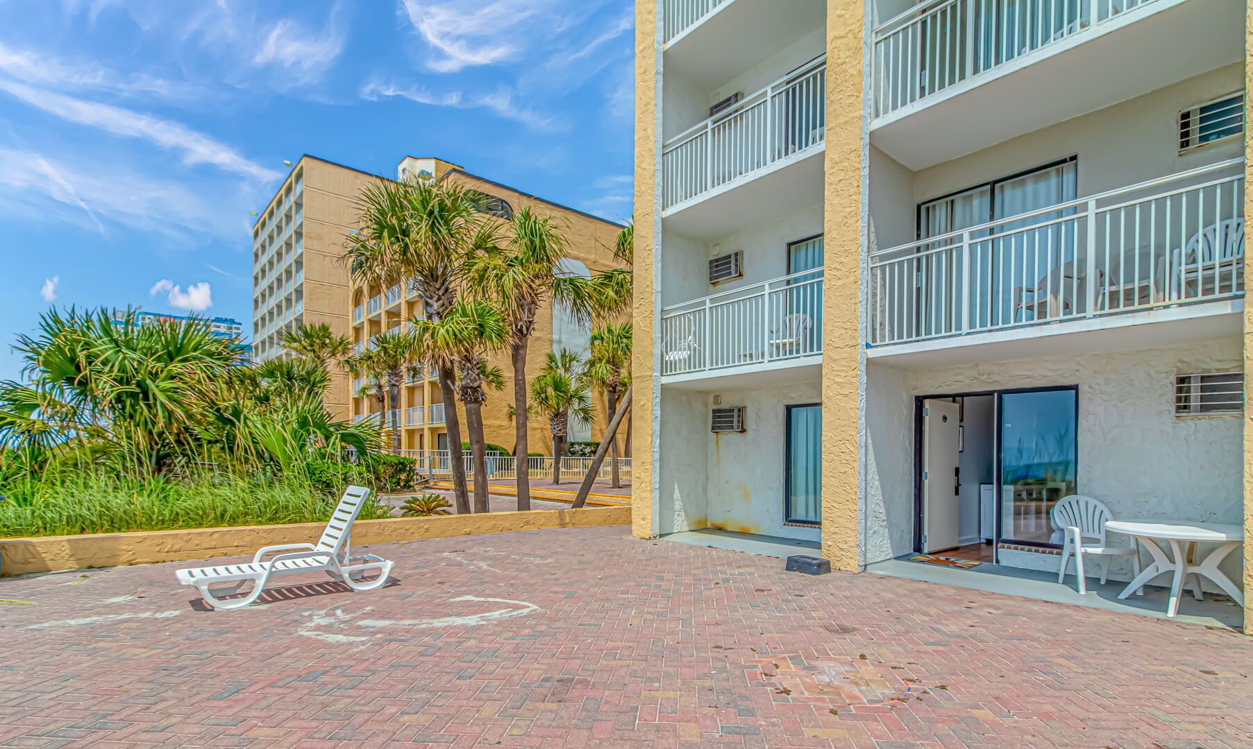 A hotel courtyard with a brick floor, palm trees, and a poolside area with lounge chairs.