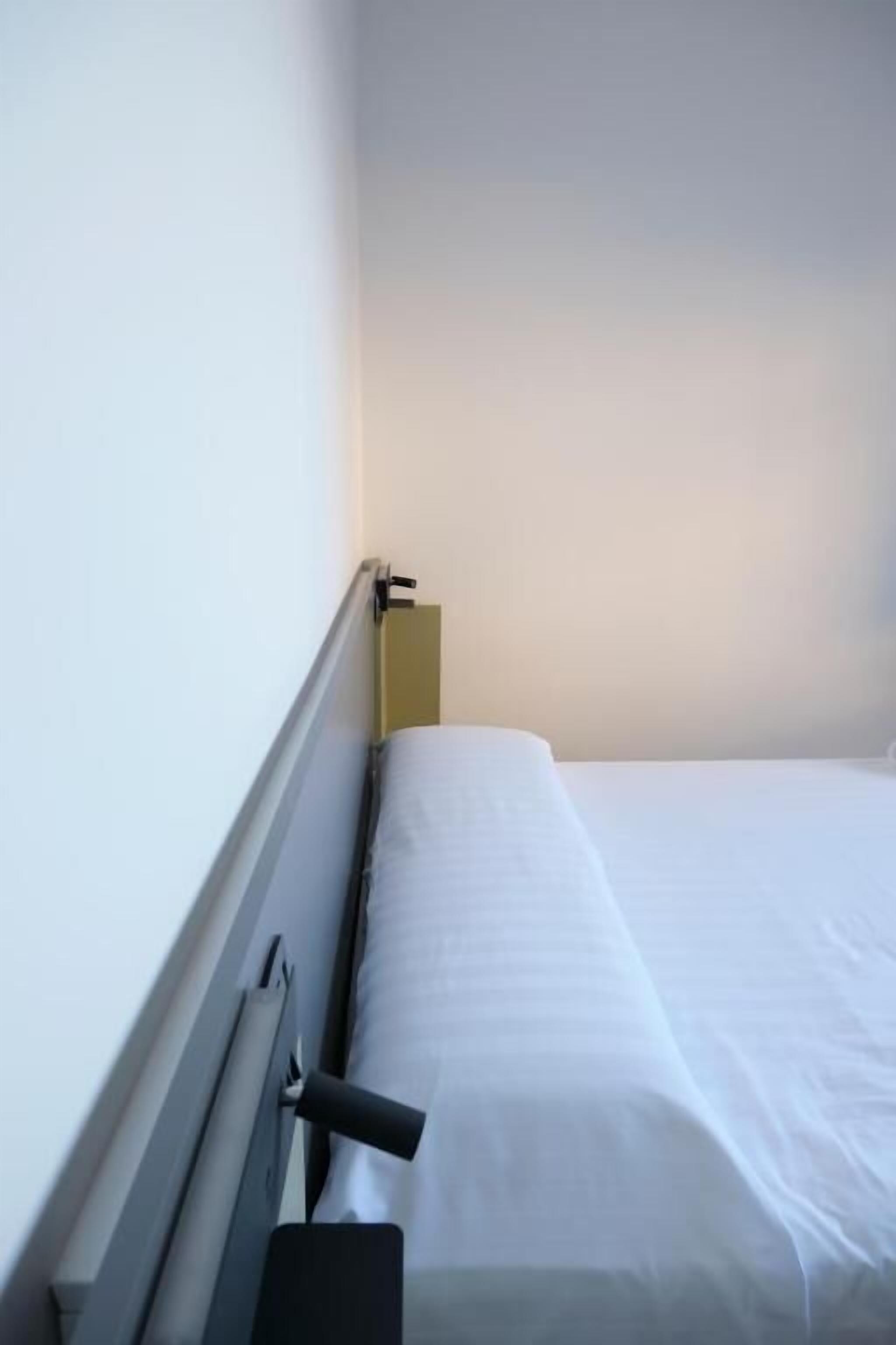 Ein Einzelbett mit weißer Bettwäsche in einem Zimmer mit schlichter Wand und einer Tür.