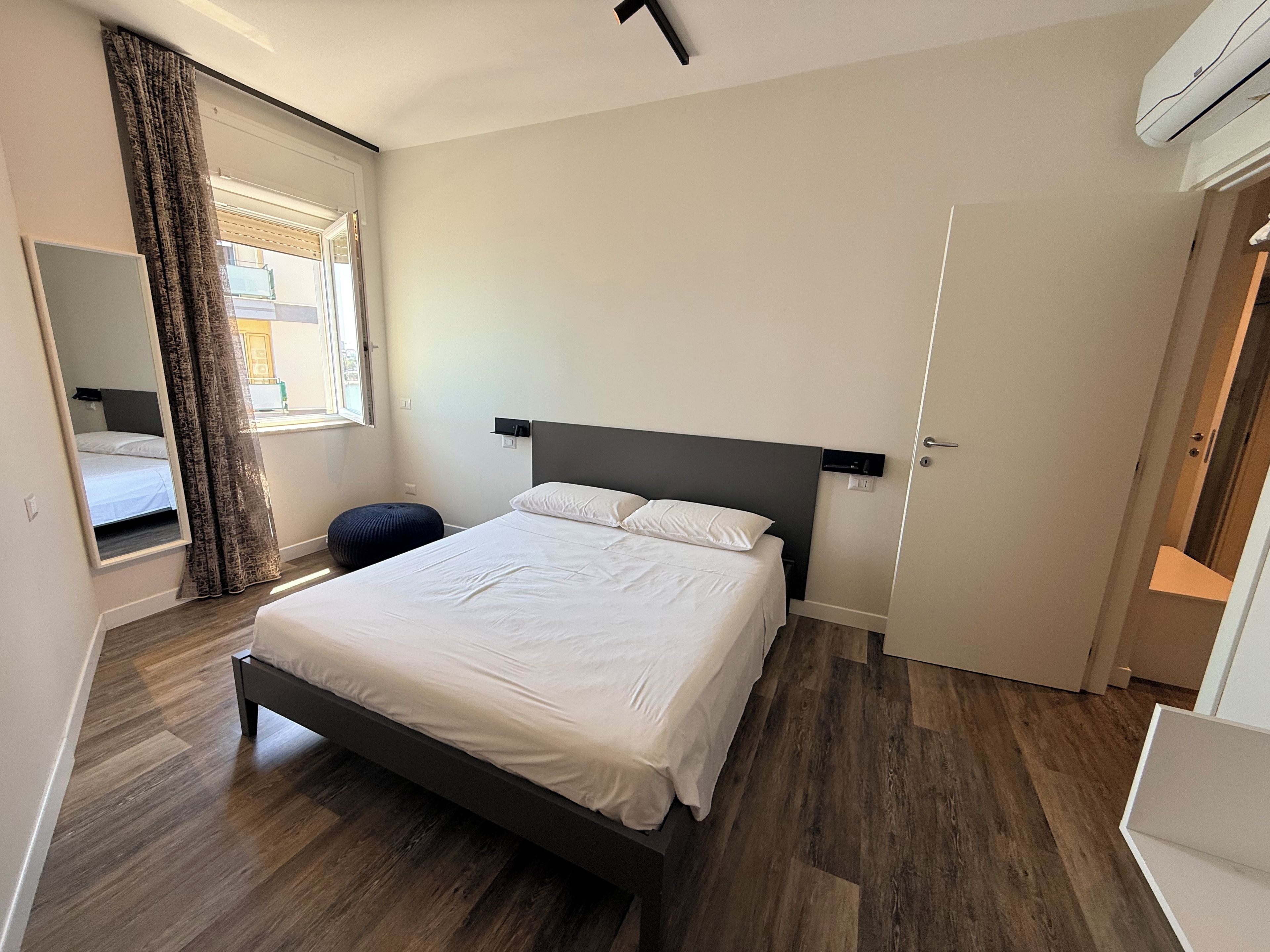A bedroom with a bed, a bedside table, a mirror, and a window with curtains.