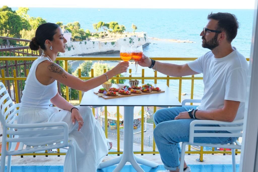 Two people toasting with drinks at a table with a view of the sea.