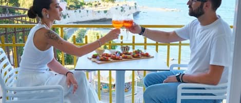 Twee personen proosten met drankjes aan een tafel met uitzicht op zee.