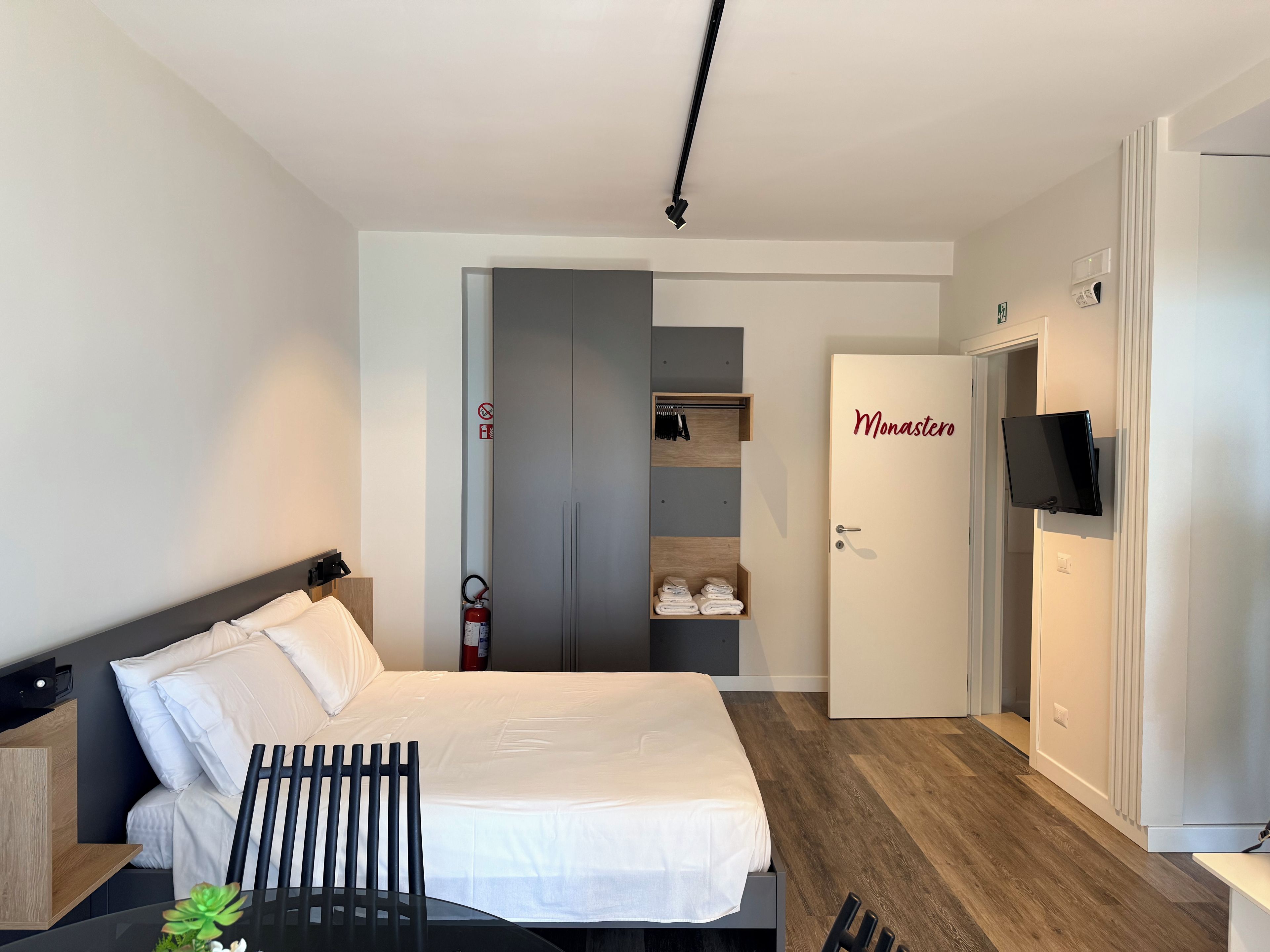 A hotel room with a bed, a desk with a chair, a television, a wardrobe, and a closet with a fire extinguisher.