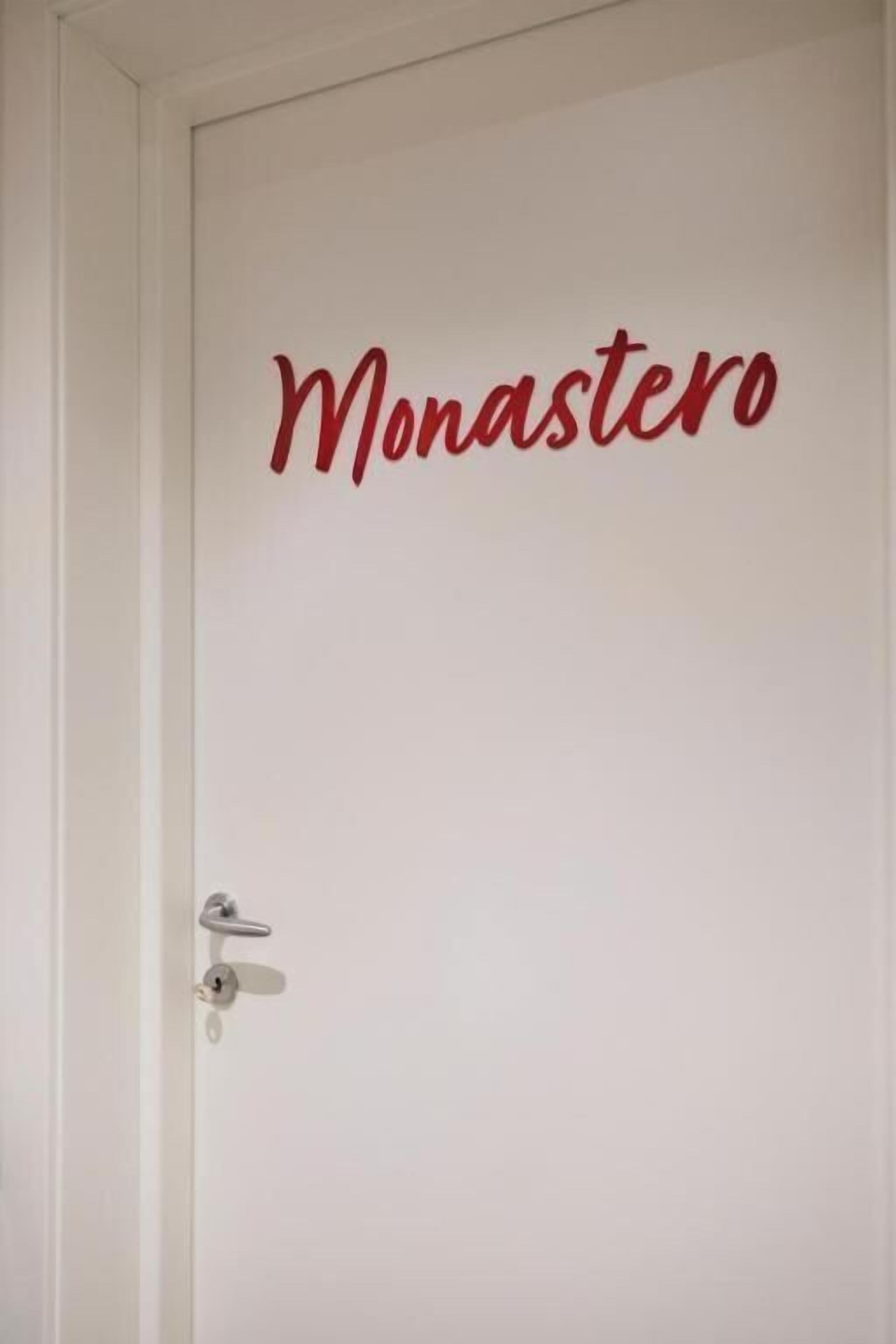 En vit dörr med ett metallhandtag och en röd skylt med texten ”Monastero” ovanför.