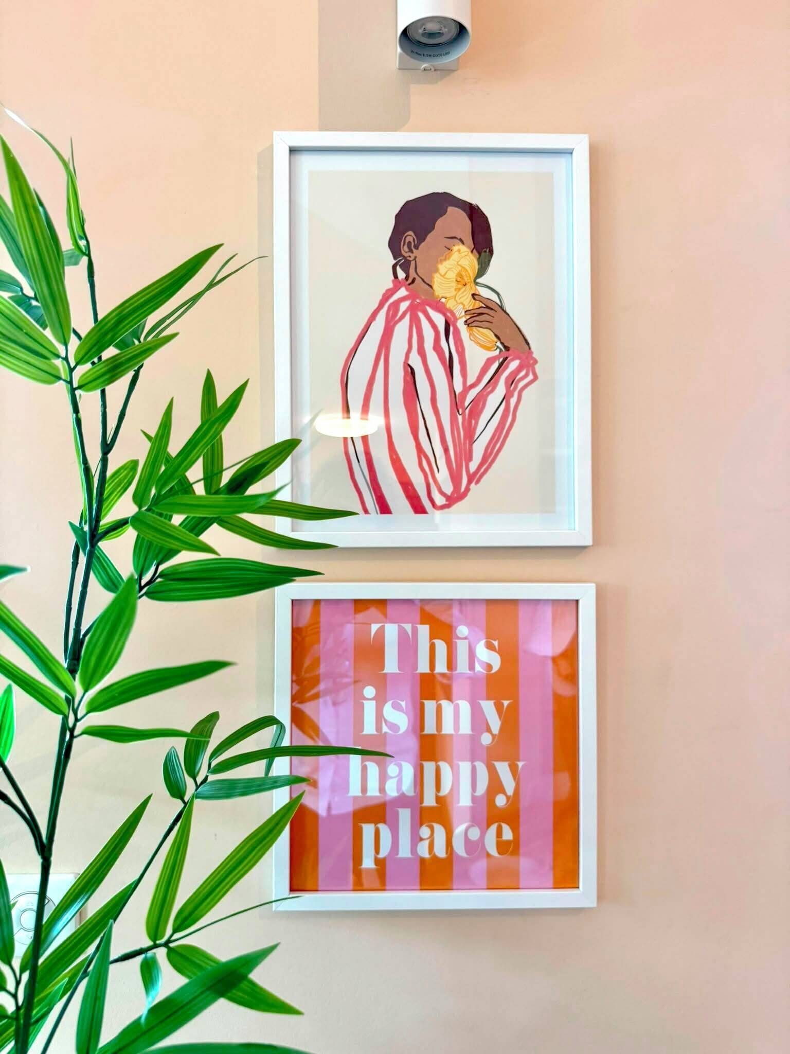 Two framed artworks on a wall, one depicting a person holding a flower and the other with the text 'This is my happy place.'