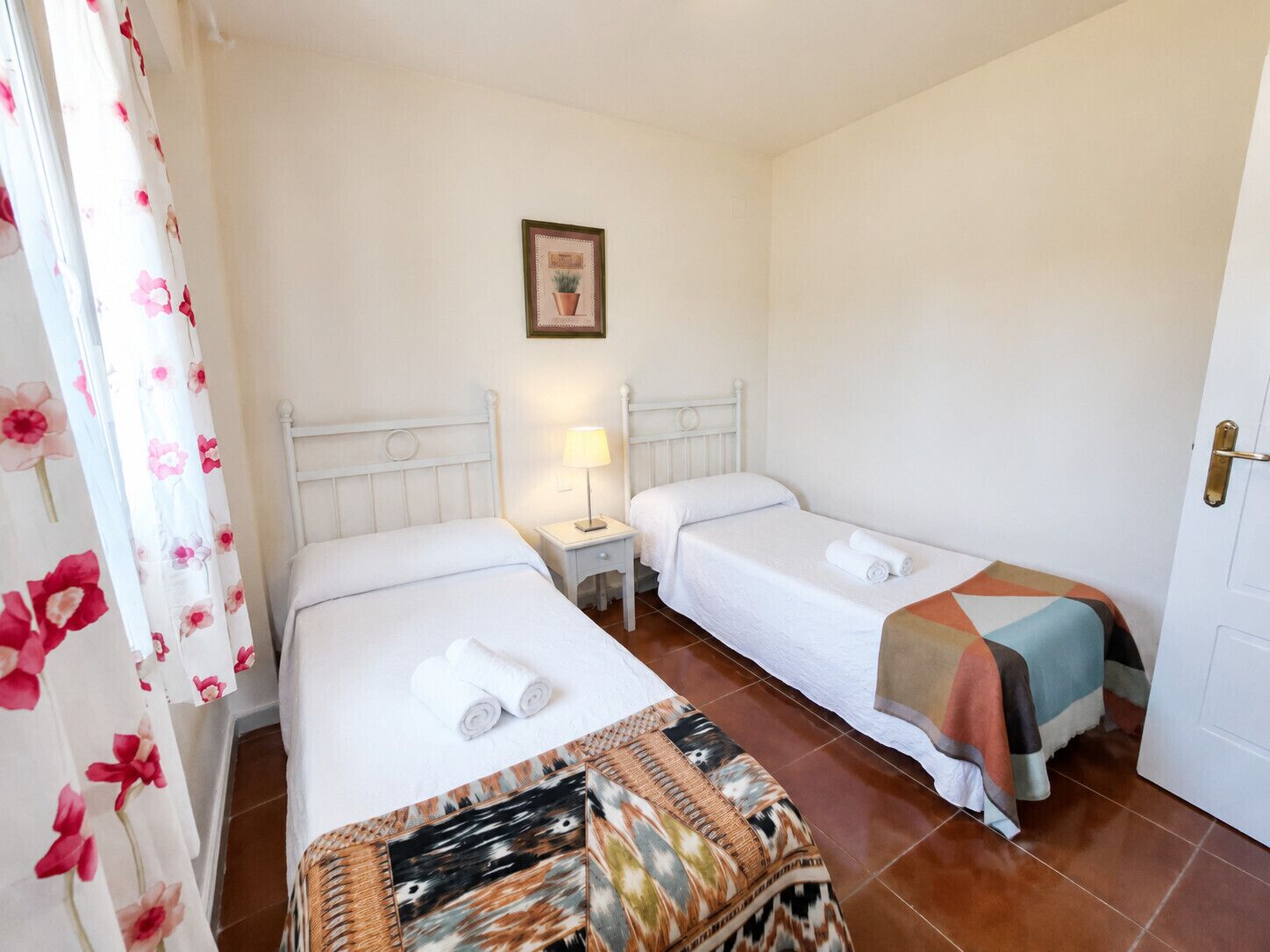 A hotel room with two single beds, a nightstand with a lamp, a framed picture on the wall, and a door with a handle.