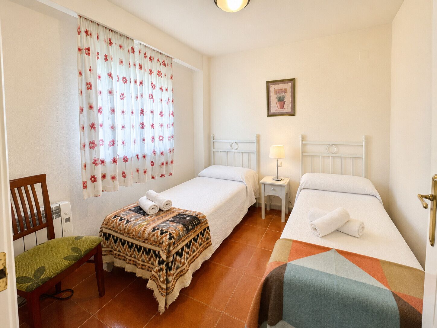 A hotel room with two beds, a chair, a bedside table with a lamp, and a window with floral curtains.