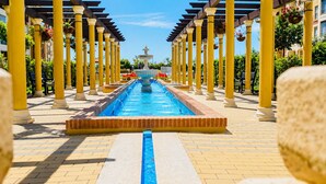 A long pool with a fountain in the center, surrounded by yellow columns and a pergola. - Los Cisnes, T2 Bajo - Ref.520B (Isla Canela)