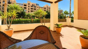 A balcony with wicker furniture, potted plants, and a view of a building with balconies. - Los Cisnes, T2 Bajo - Ref.520B (Isla Canela)