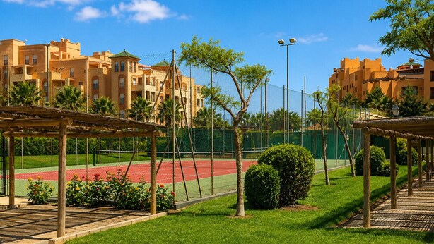 A tennis court surrounded by a fence, with a building in the background. - Los Cisnes, T2 Bajo - Ref.520B (Isla Canela)