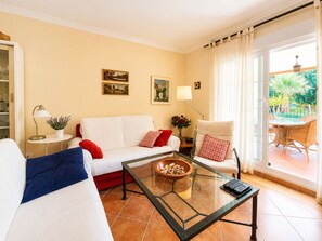 Ein Wohnzimmer mit Sofa, Sessel, Glastisch und Blick auf eine Terrasse.