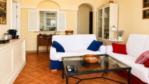 A living room with a white sofa, a glass coffee table, and a kitchen area in the background. - Los Cisnes, T2 Bajo - Ref.520B (Isla Canela)