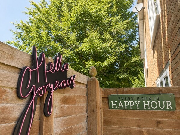 A wooden fence with a neon sign that reads 'Hello Gorgeous' and a green sign stating 'Every Hour is Happy Hour.'