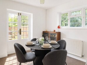 A dining area with a round table set for four, surrounded by four chairs. There are windows on one side and a radiator on the other.