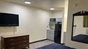 Quarto de hotel com cama, cômoda, televisão, cozinha compacta com micro-ondas e geladeira, e espelho.