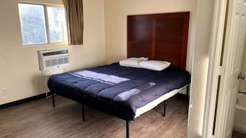 Uma cama de solteiro com cabeceira escura, uma janela com cortinas e um ar condicionado fixado na parede.