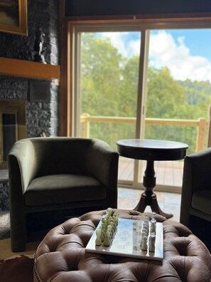 Ein Zimmer mit Kamin, einem Schachbrett auf einem Ledersitzhocker und Blick auf eine Terrasse sowie Bäume durch ein großes Fenster.