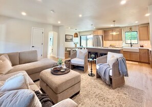 Ein modernes Wohnzimmer mit Sofa, Hocker und Sesseln, eine Küche mit Holzschränken und Edelstahlgeräten sowie ein Essbereich mit Tisch und Stühlen.