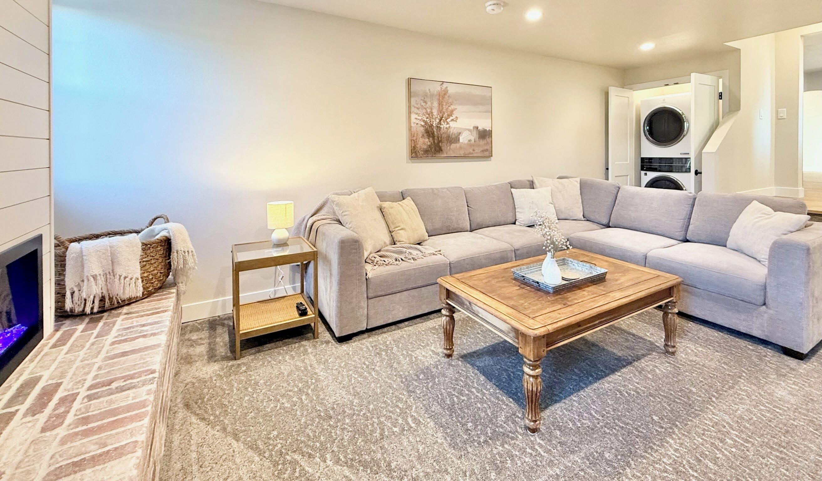A living room with a sectional sofa, a wooden coffee table, a side table with a lamp, a fireplace, and a washing machine in the background.