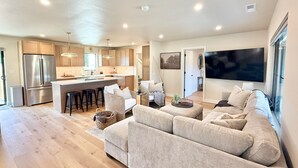 A modern kitchen with a central island, a dining area with a sofa and chairs, and a large flat-screen TV mounted on the wall. - The Ayres House North Gilham-UofO-Autzen-Oakway, Putting Green Large Turfed Yard (Eugene)
