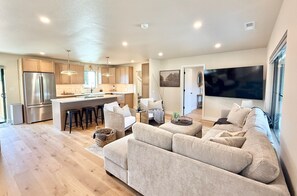 A modern kitchen with a central island, a dining area with a sofa and chairs, and a large flat-screen TV mounted on the wall. - The Ayres House North Gilham-UofO-Autzen-Oakway, Putting Green Large Turfed Yard (Eugene)