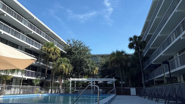 Zwembadgebied van een hotel, met een helderblauwe lucht, palmbomen en een meerlaags gebouw met balkons.