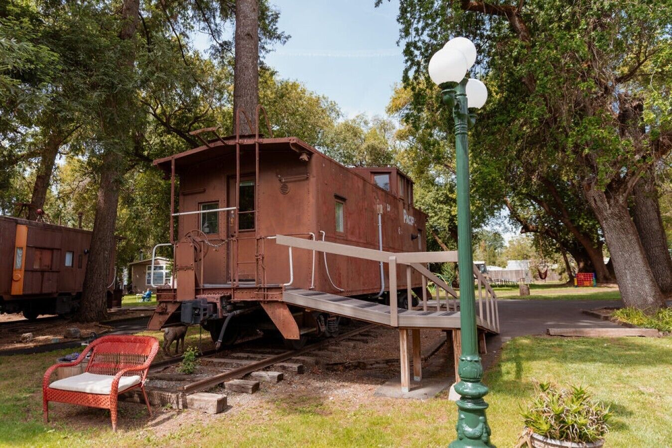 A stationary train caboose with a small platform, surrounded by trees and a red chair.