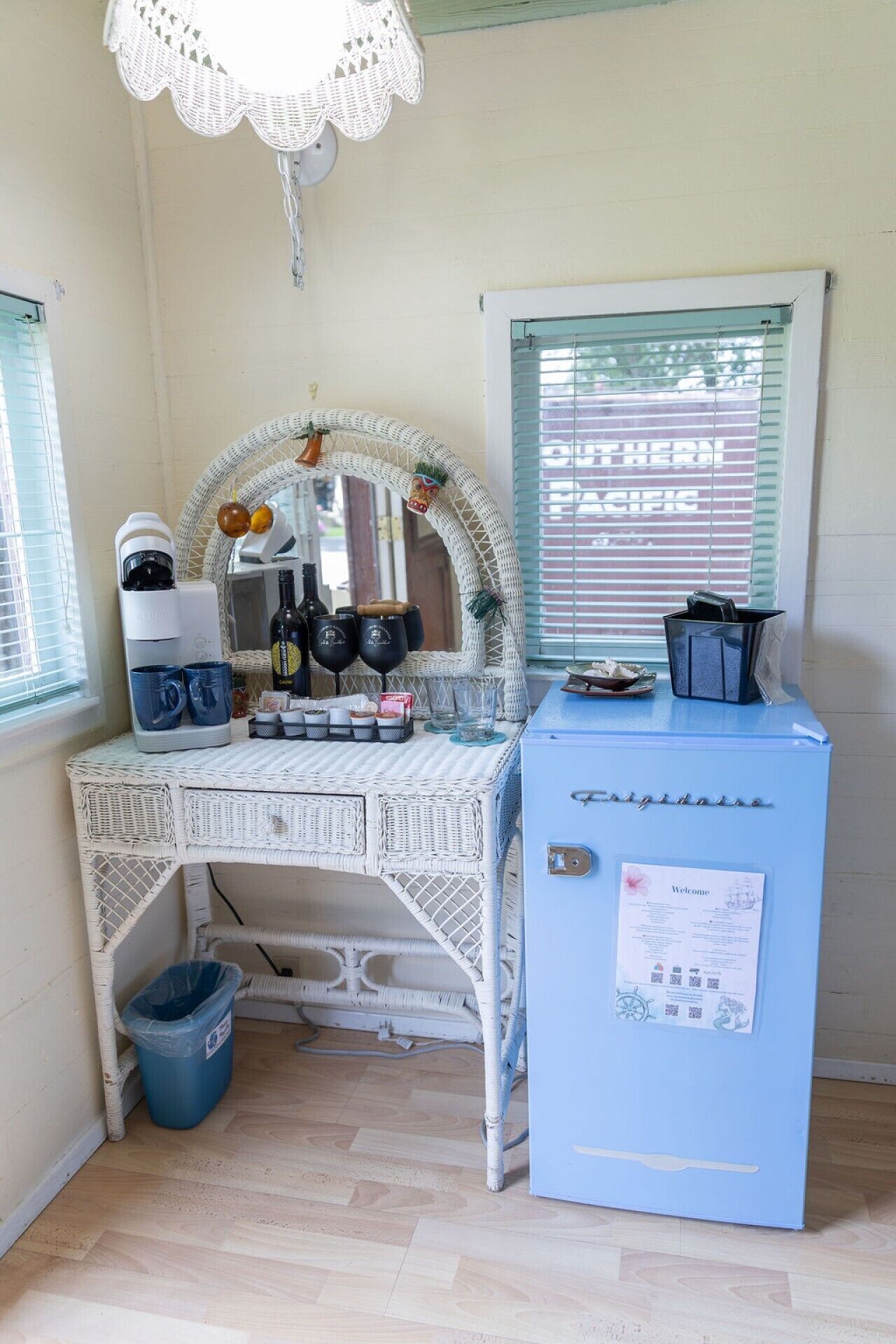 A room with a white wicker desk, a coffee maker, mugs, and a blue cabinet with a welcome sign.